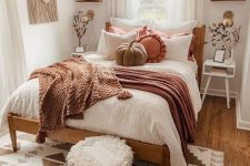 a narrow boho bedroom with a stained bed and muted color bedding, white nightstands, a fringe pouf, boho textiles and some fall decor