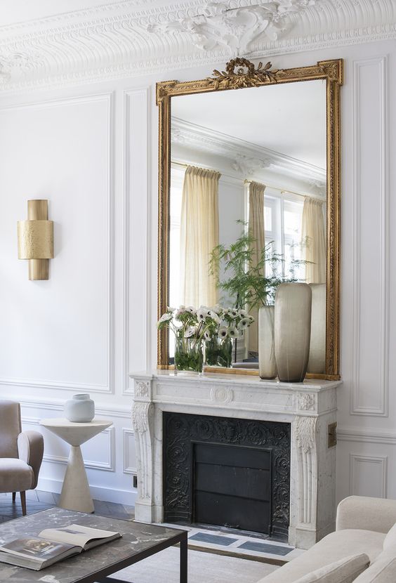 a neutral Parisian living room with a fireplace and a stone mantel, an oversized mirror in a gold frame, blooms and neutral seating furniture