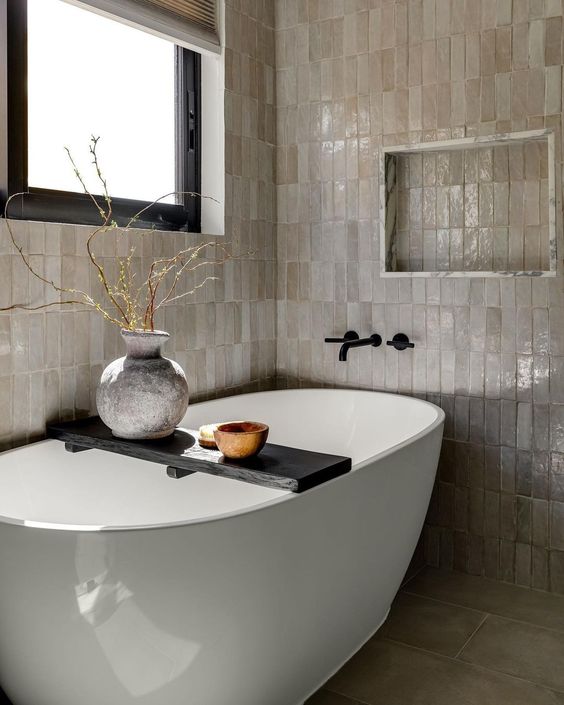 a neutral bathroom with stacked Zellige tiles, an oval tub, a niche, some branches in a vase and a window