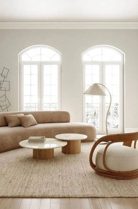 a neutral living room with a curved tan sofa, round coffee tables, a curved rattan and boucle chair, floor lamp and a neutral rug