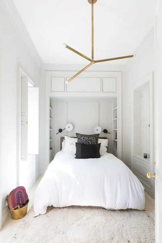 A neutral narrow bedroom with built in shelves at the headboard, a bed with black and white bedding, a chandelier and some bright decor