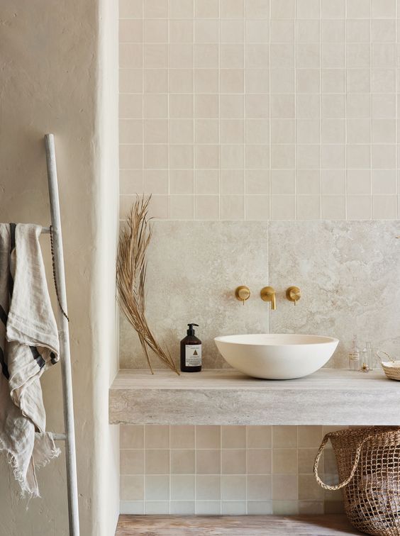 a neutral organic bathroom clad with tan Zellige and stone tiles, a stone vanity, a sink and brass fixtures