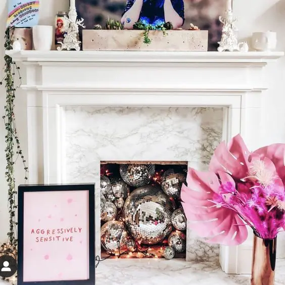 A non working fireplace filled with disco balls, with a mantel with greenery, a vase with pink dried leaves and a sign