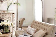 a refined French living room with a carved fireplace, a large floor mirror, a neutral loveseat, a table with blooms and a chic chandelier