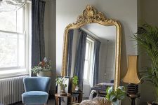 a refined Victorian bedroom with dove grey walls, a bed, a periwinkle bench, a blue chair, a mirror in a gold ornated frame and potted greenery