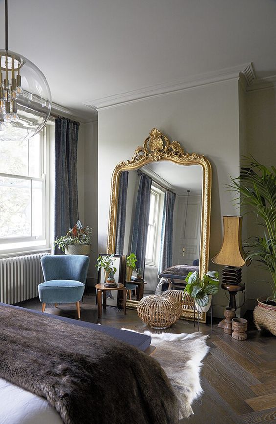 a refined Victorian bedroom with dove grey walls, a bed, a periwinkle bench, a blue chair, a mirror in a gold ornated frame and potted greenery