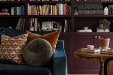 a refined and moody space with aubergine arched bookcases, a navy sofa with pillows, a vintage table and a green chair