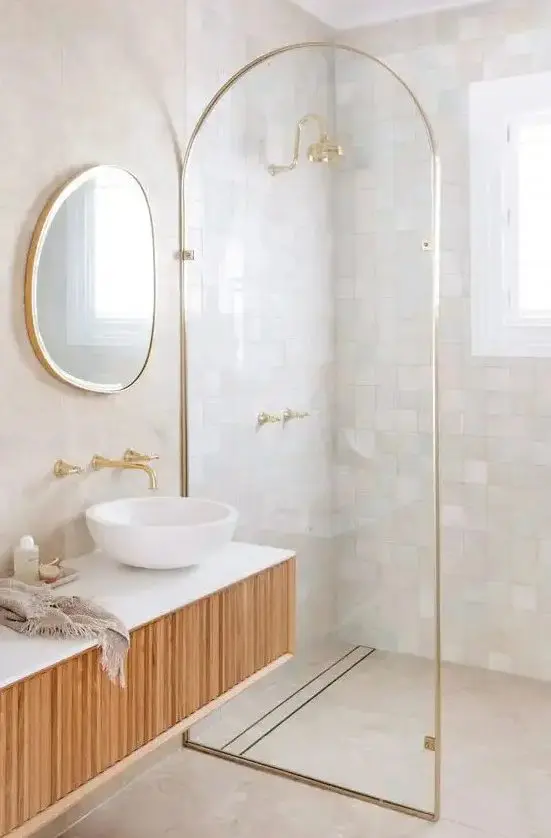 a refined bathroom clad with neutral and pastel zellige tiles in the shower, with a curved space divider and a floating wooden vanity