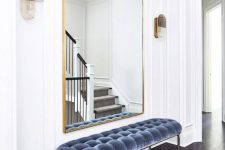 a refined minimalist entryway with a pretty arched mirror in a gilded frame, a blue uphalstered bench and sconces