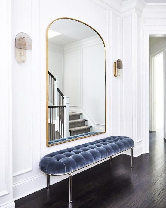 a refined minimalist entryway with a pretty arched mirror in a gilded frame, a blue uphalstered bench and sconces