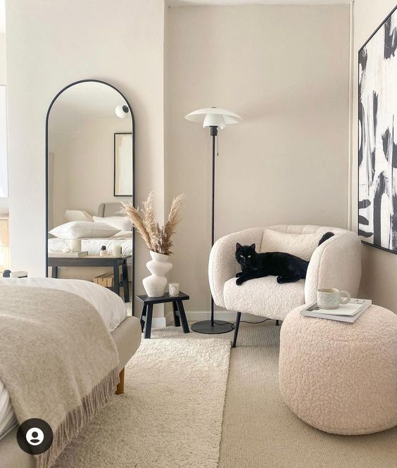 a refined neutral bedroom with a grey bed and neutral bedding, a white boucle chair and a pouf, a black side table and an arched mirror
