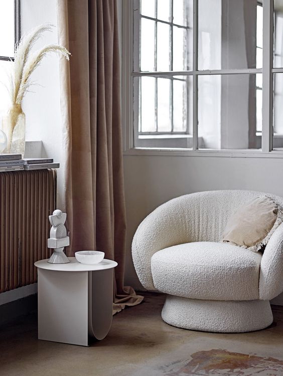 a refined nook with a white boucle chair, a pillow, a side table, some lovely decor and beige curtains