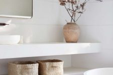 a serene white bathroom done with terracotta herringbone tile, an open vanity with baskets, a tub and some dried branches
