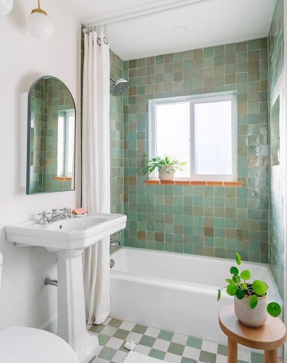 A small and cute bathroom with green and beige Zellige tiles, a checked floor, a free standing tub, some potted plants