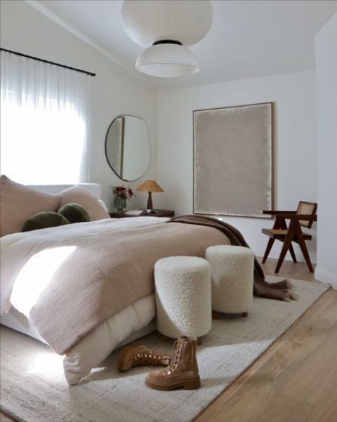 a small and lovely bedroom with a white bed and pastel bedding, white boucle poufs, a stained chair and a neutral rug