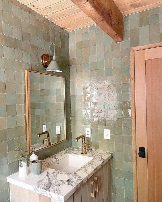 a small bathroom done with green and beige Zellige tiles, a neutral vanity, a mirror and brass fixtures