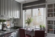 a small grey kitchen with dark countertops and a windowsill as a table, tall burgundy chairs and lights over this zone