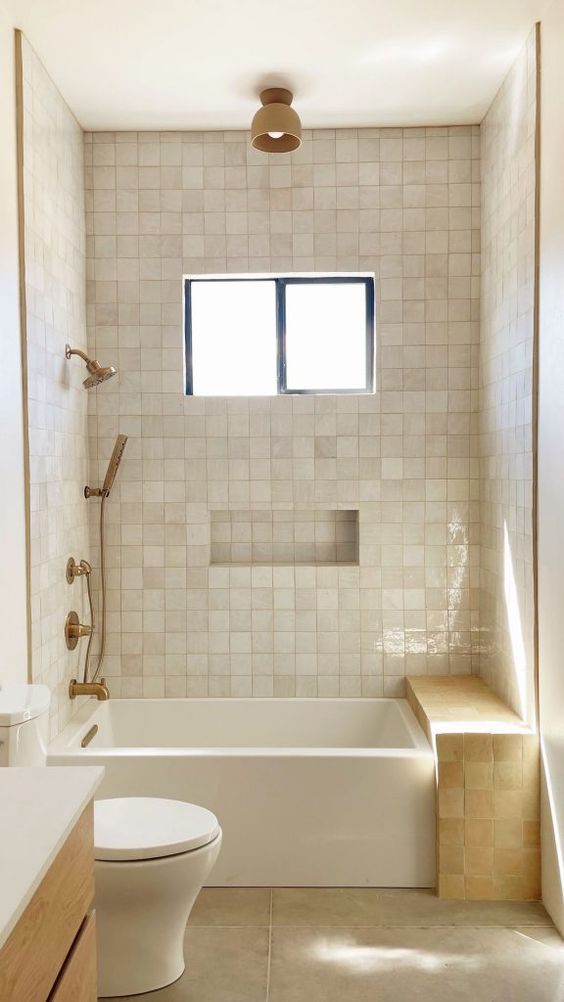 a small neutral bathroom clad with Zellige tiles, with a tub and a Zellige tile bench, brass fixtures and a window
