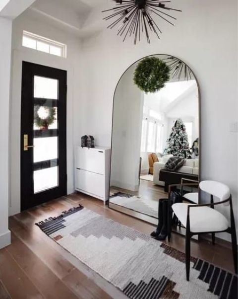 A small yet eye catchy entryway with a large arched mirror, a whiet shoes cabinet, a rug and a chair