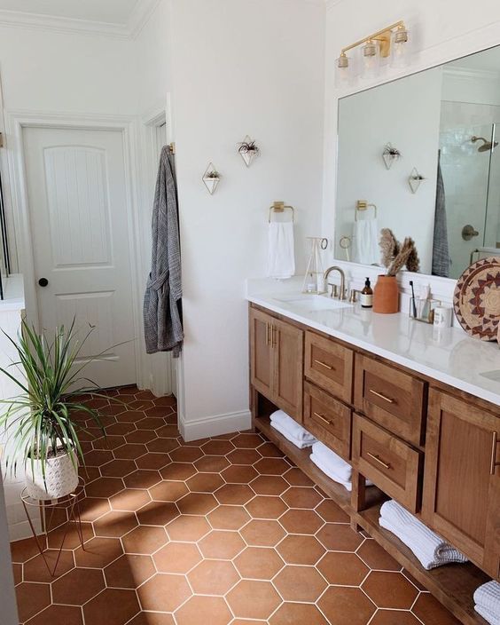 a stylish and cozy bathroom with white walls and a terracotta hex tile floor, a stained vanity, greenery and a large mirror