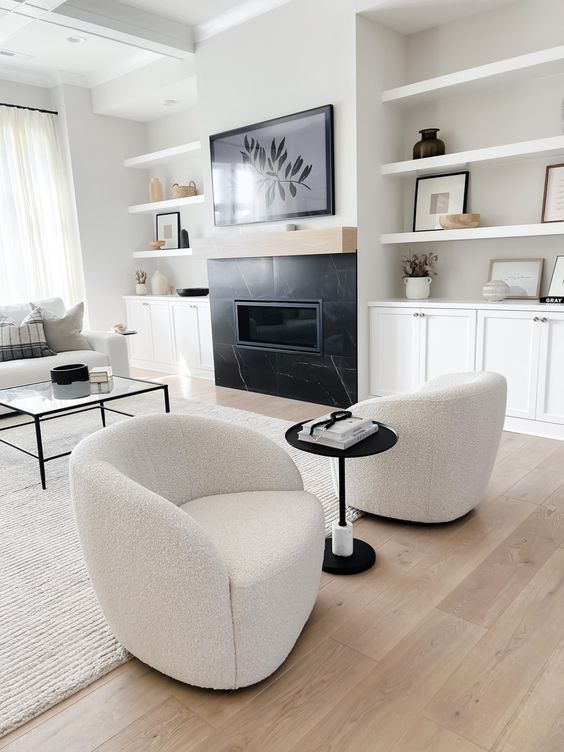 A stylish living room with a built in fireplace, built in cabinets and shelves, a glass coffee table, white boucle chairs and a sofa