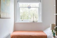 a tiny bedroom with a bed with a rust blanket, a box with blankets, a shelving unit and some decor will do as a guest bedroom or even a kid’s room