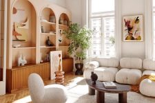 a trendy living room with peachy pink arched bookcases and stained cabinets, a white low sofa and curved chairs, a coffee table