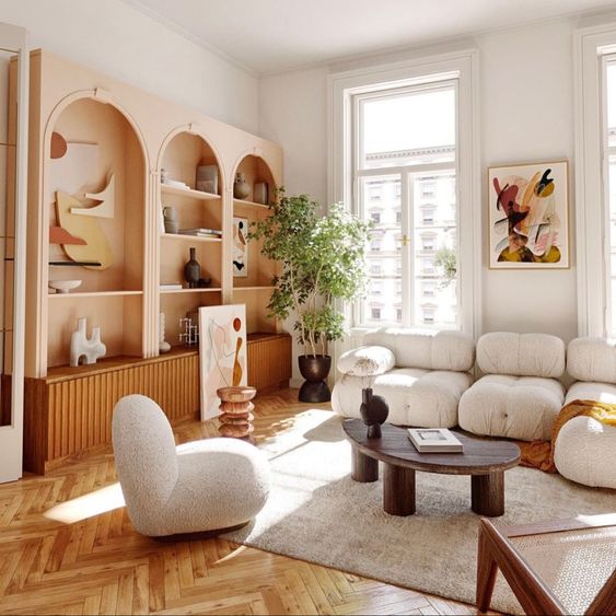 a trendy living room with peachy pink arched bookcases and stained cabinets, a white low sofa and curved chairs, a coffee table