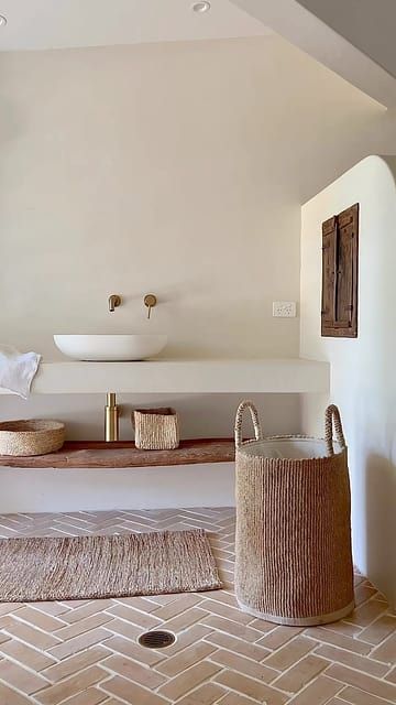 A wabi sabi meets Mediterranean bathroom in white, with a herringbone terracotta floor, an open vanity and a basket