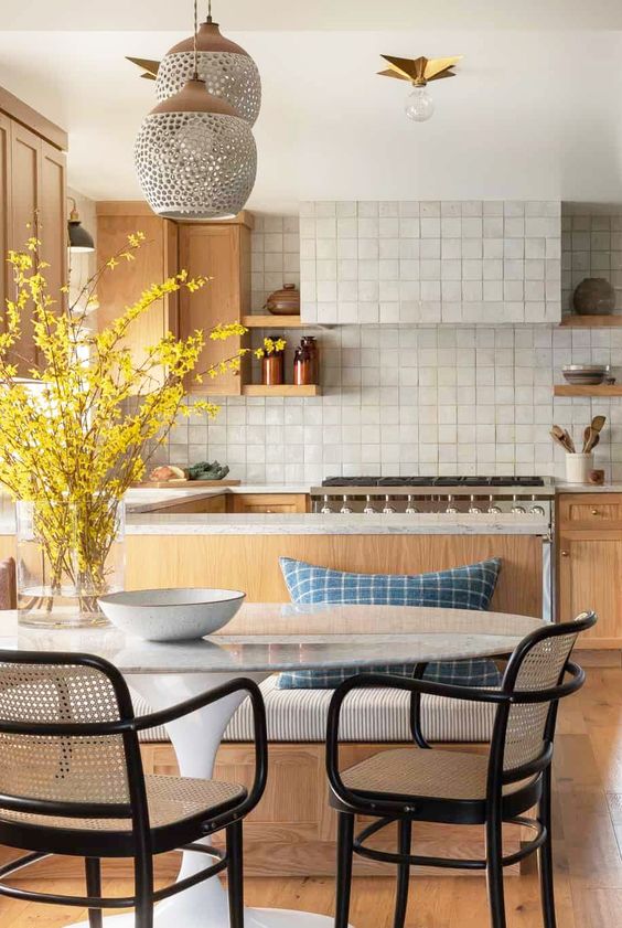 A welcoming kitchen with light stained cabinetry, a white Zellige tile bacskplash, chic dining furniture