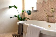 a whimsical and chic earthy bathroom clad with herringbone terracotta tiles, a large tub, a potted plant, gold fixtures