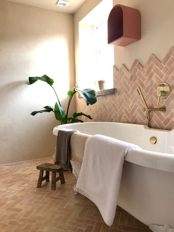 a whimsical and chic earthy bathroom clad with herringbone terracotta tiles, a large tub, a potted plant, gold fixtures