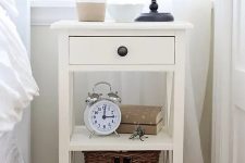 a white cottage-style nightstand with a basket, a drawer and some decor and a black metal lamp