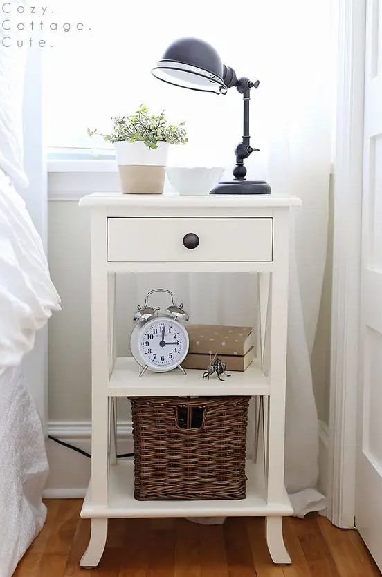 A white cottage style nightstand with a basket, a drawer and some decor and a black metal lamp