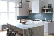 a white kitchen with a matte blue stacked tile backsplash, a neutral kitchen island with a stone countertop and a chandelier