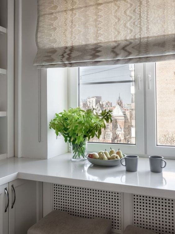 a white kitchen with countertops, one of which doubles as a dining table, with a printed curtain and a view