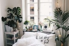 a white narrow bedroom with a small bed, a nightstand with storage, potted plants, decor and lights over the bed