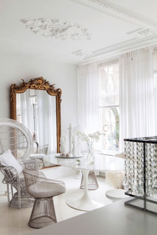 An all white dining room with a mirror in an ornated frame, a white table and metal and papasan chairs is wow