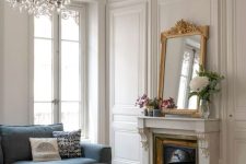 an elegant Parisian living room with chevron floors, molding, a fireplace with a large mirror, a navy sofa, some tables and a crystal chandelier