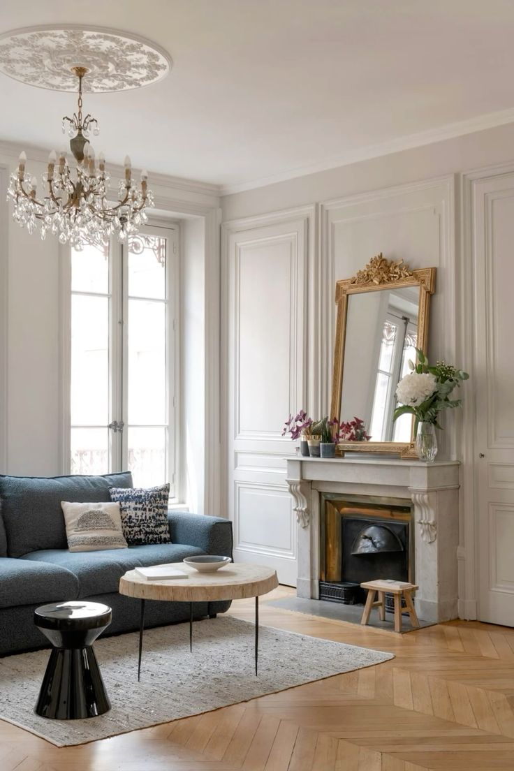 an elegant Parisian living room with chevron floors, molding, a fireplace with a large mirror, a navy sofa, some tables and a crystal chandelier