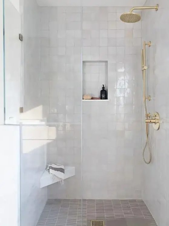 an elegant white shower space completely clad with white zellige tiles, with gold fixtures and a small niche for storage