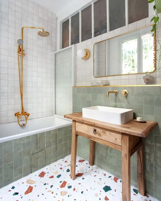 An eye catchy bathroom clad with white and green Zellige tiles, a terrazzo floor, a stained vanity and gold fixtures
