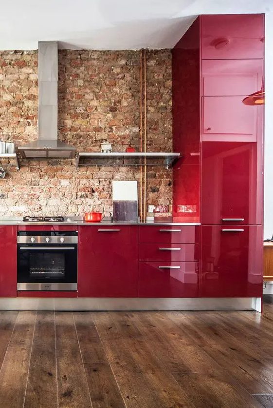 an eye-catchy deep red glossy ktchen with a red brick backsplash, grey countertops and stainless steel appliances