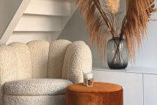 an under the stairs nook with a white scallop boucle chair, a rust-colored side table, a white storage unit and pampas grass
