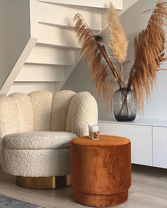 An under the stairs nook with a white scallop boucle chair, a rust colored side table, a white storage unit and pampas grass