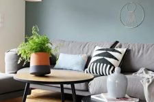 a Scandinavian living room with a green grey accent wall, a grey sofa and a chair, a couple of coffee tables and orange decor