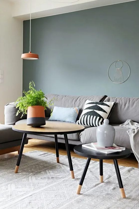 a Scandinavian living room with a green grey accent wall, a grey sofa and a chair, a couple of coffee tables and orange decor