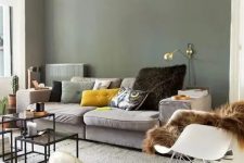 a Scandinavian living room with olive green walls, a grey sofa and colorful pillows, a gold sconce, a rocker and some coffee tables