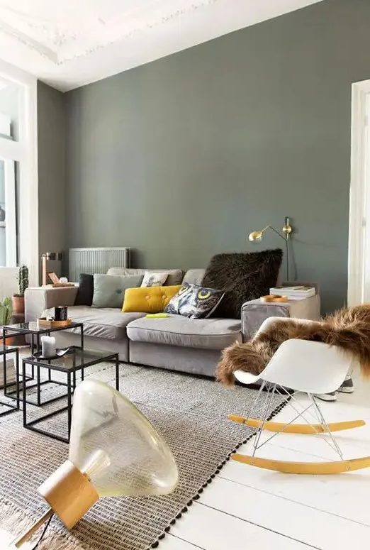 a Scandinavian living room with olive green walls, a grey sofa and colorful pillows, a gold sconce, a rocker and some coffee tables