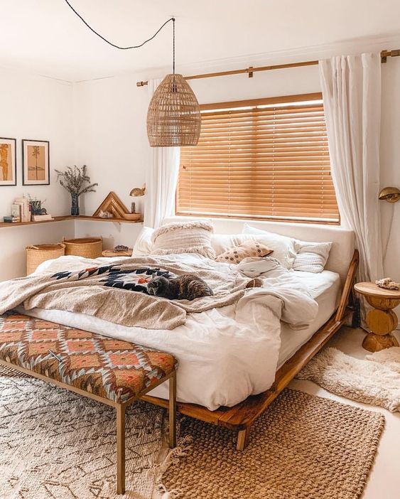 a boho bedroom with layered rugs, a bed with neutral bedding, a boho bench, baskets, a nightstand, a pendant lamp and cool decor
